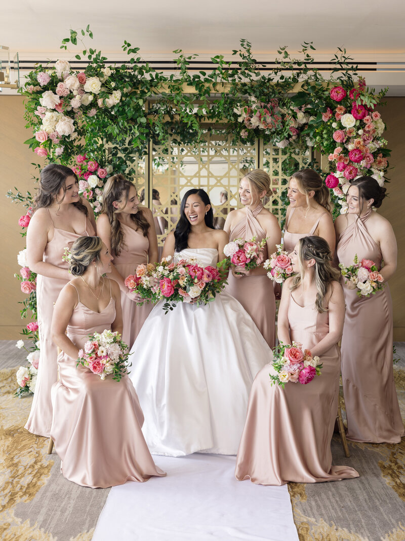 A bride in a white gown sits in front of a floral archway, surrounded by seven bridesmaids in matching pink dresses, all holding bouquets of pink and white flowers. They are smiling and joyful.