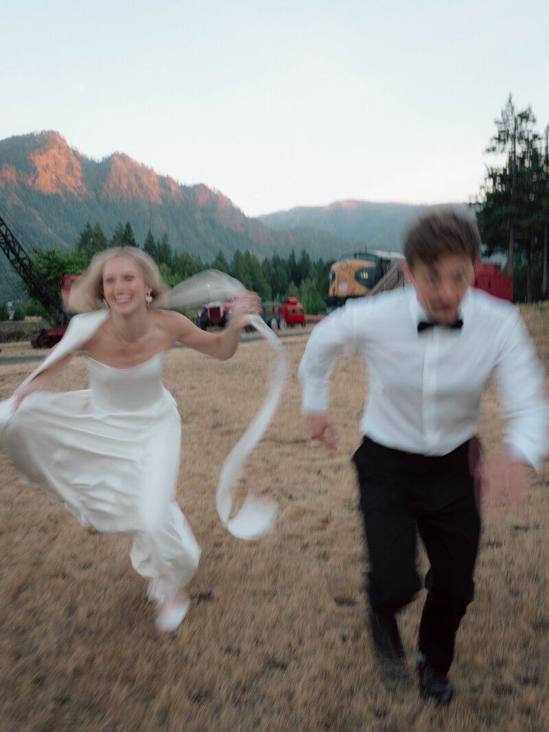Bride and groom running joyfully through field at sunset with mountain backdrop