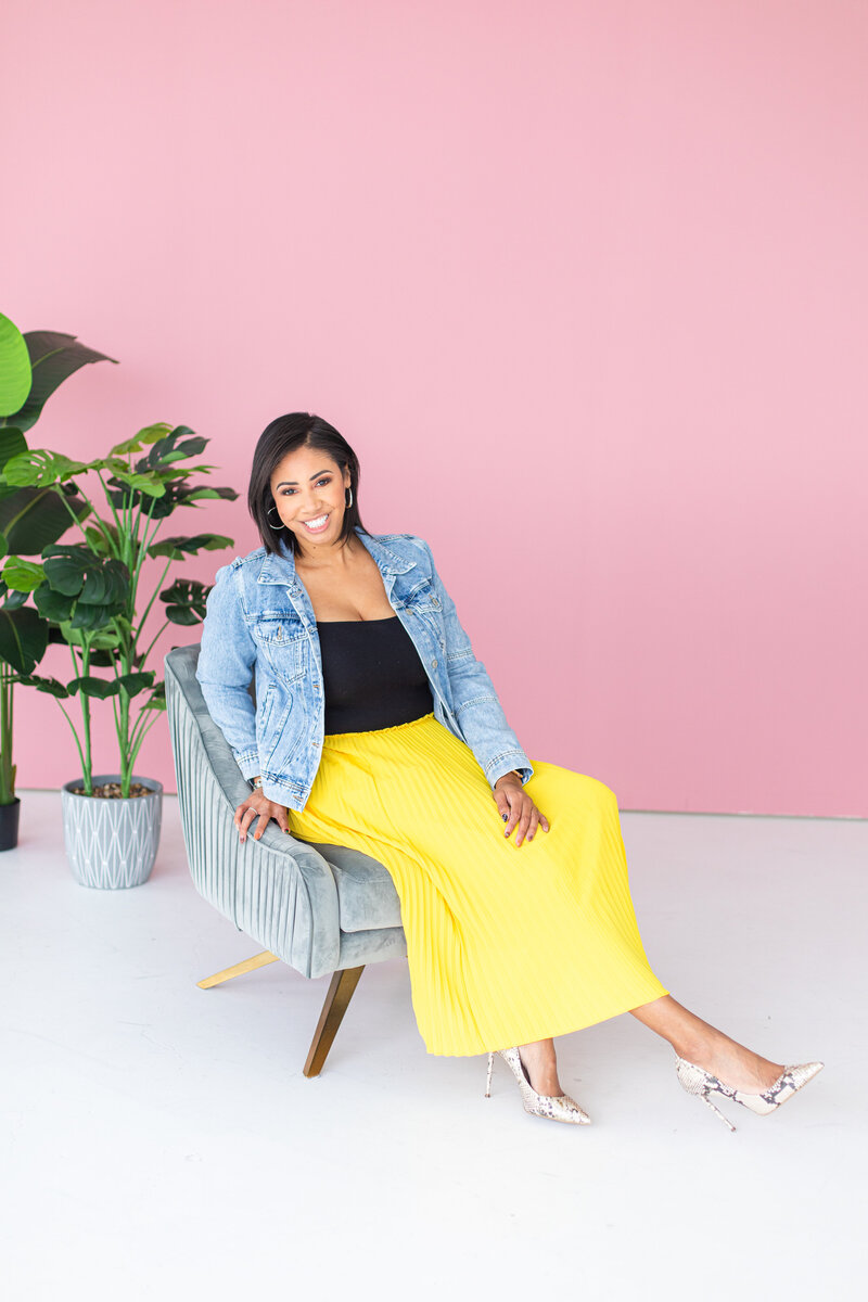 Sheena is sitting on a grey  and brown couch. she wears a jean jacket, black tank top, yellow skirt and elegant heels. she is in front of a pink wall and plants