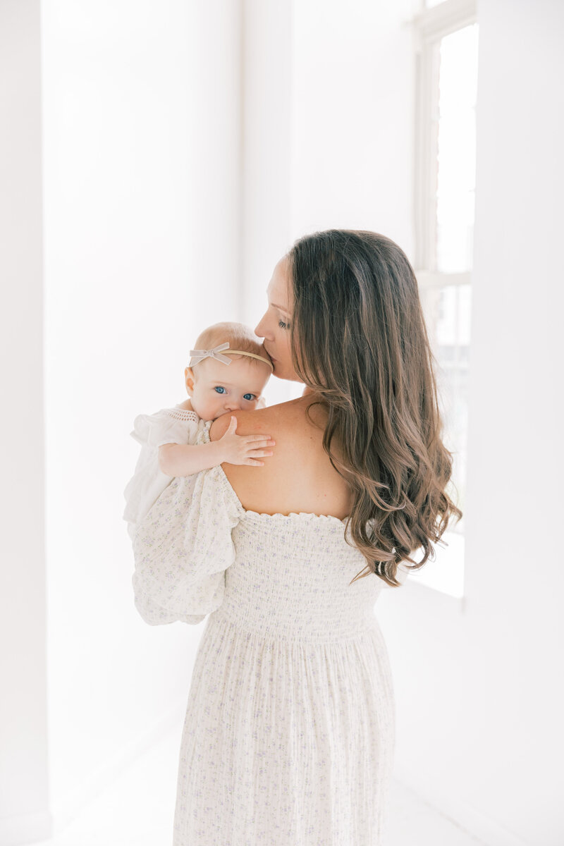 A Mother kisses the head of her infant daughter resting on her shoulder while standing