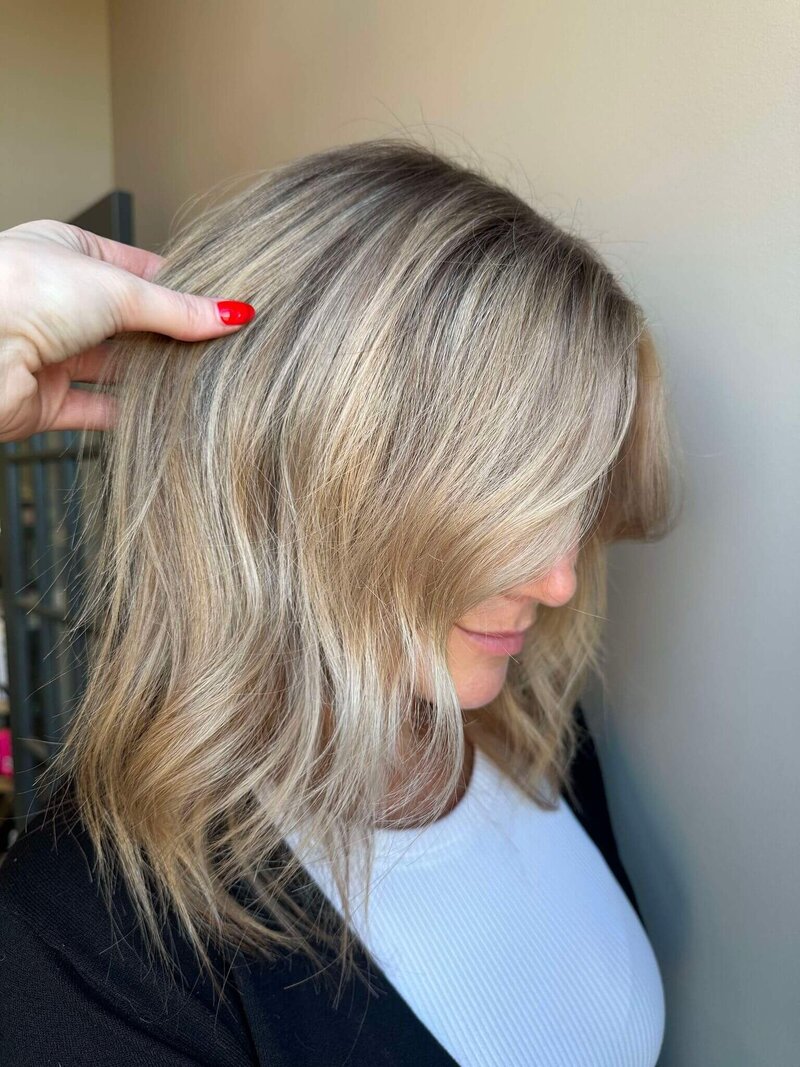 A woman with a chic, wavy blonde bob hairstyle, with a hand gently adjusting her hair, showcasing a stylish cut created by 212 Salon, Spa, & Barbershop.