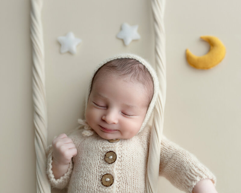 Baby boy posed on a swing with celestial moon-and-star props, dressed in a cozy cream knit outfit—dreamy and gentle.