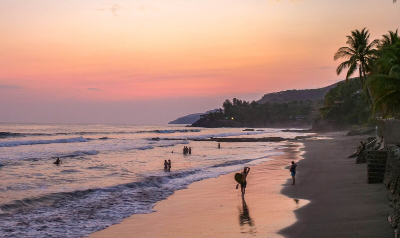 A serene beach at sunset with the sky glowing in shades of pink and orange. Gentle waves roll onto the shore where several people are swimming and wading in the water. A surfer carrying a board walks along the wet sand, leaving reflections behind, while another person strolls nearby. 
