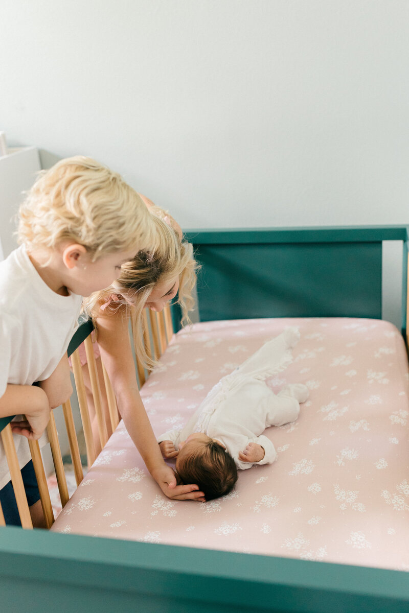 Excited older siblings lean over new baby sisters crib during in home newborn photos in Tampa, Florida