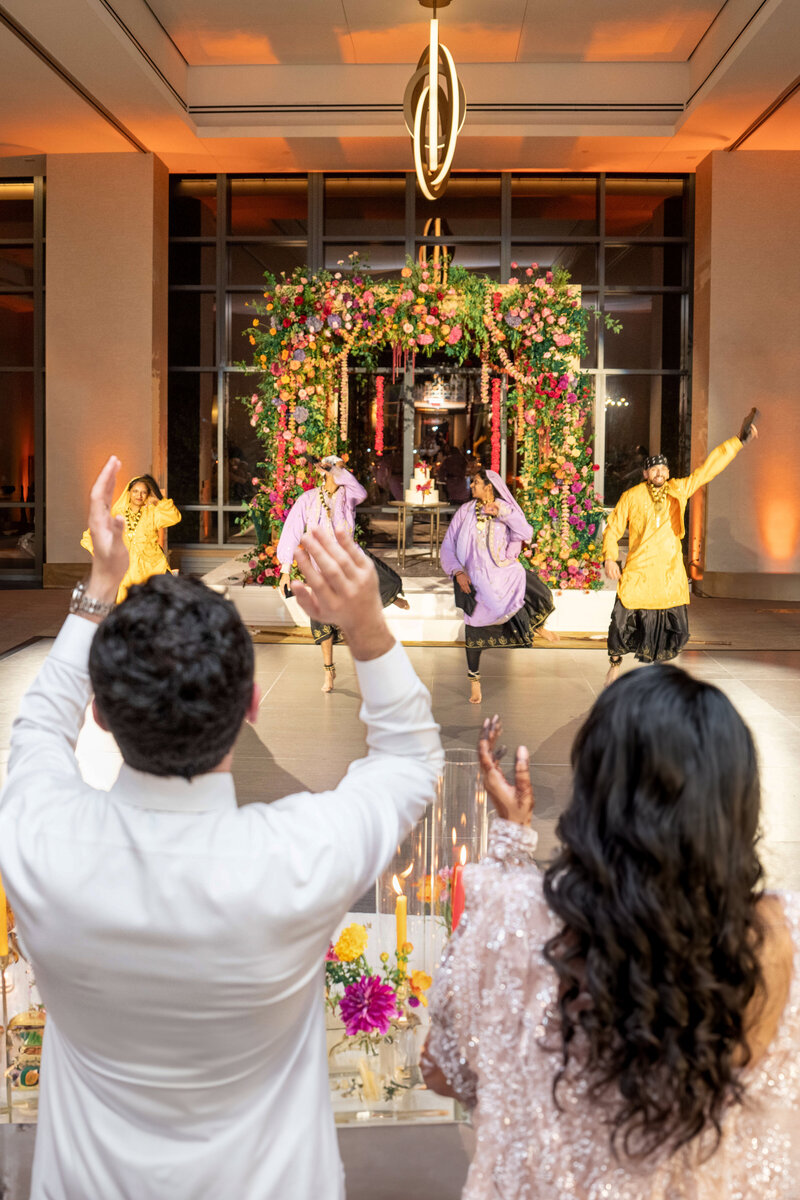 A couple claps while watching a colorful dance performance. Four dancers in vibrant costumes perform energetically on a decorated stage with flowers. The room is elegantly lit, creating a festive atmosphere.