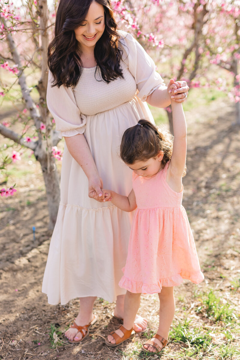 mom in ivory dress holding daughter's hands while dancing in flowering orchard