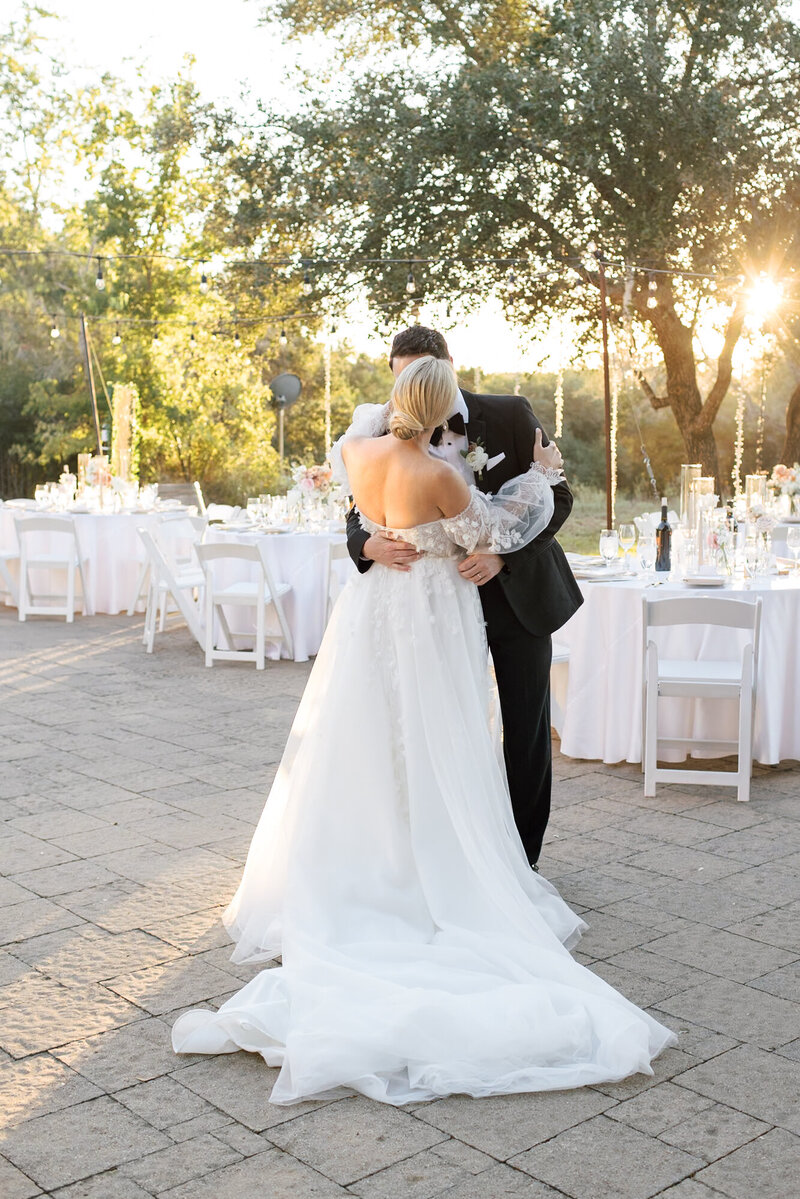 Bride and groom kissing in front of white table and chairs, sun setting in background