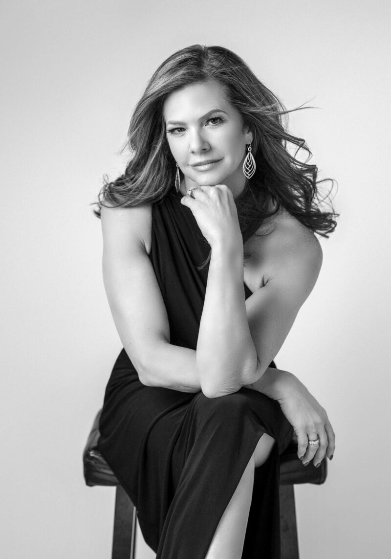 A striking black-and-white portrait of a woman in a sleek black dress, sitting with her hand gently resting on her chin, looking poised and sophisticated.