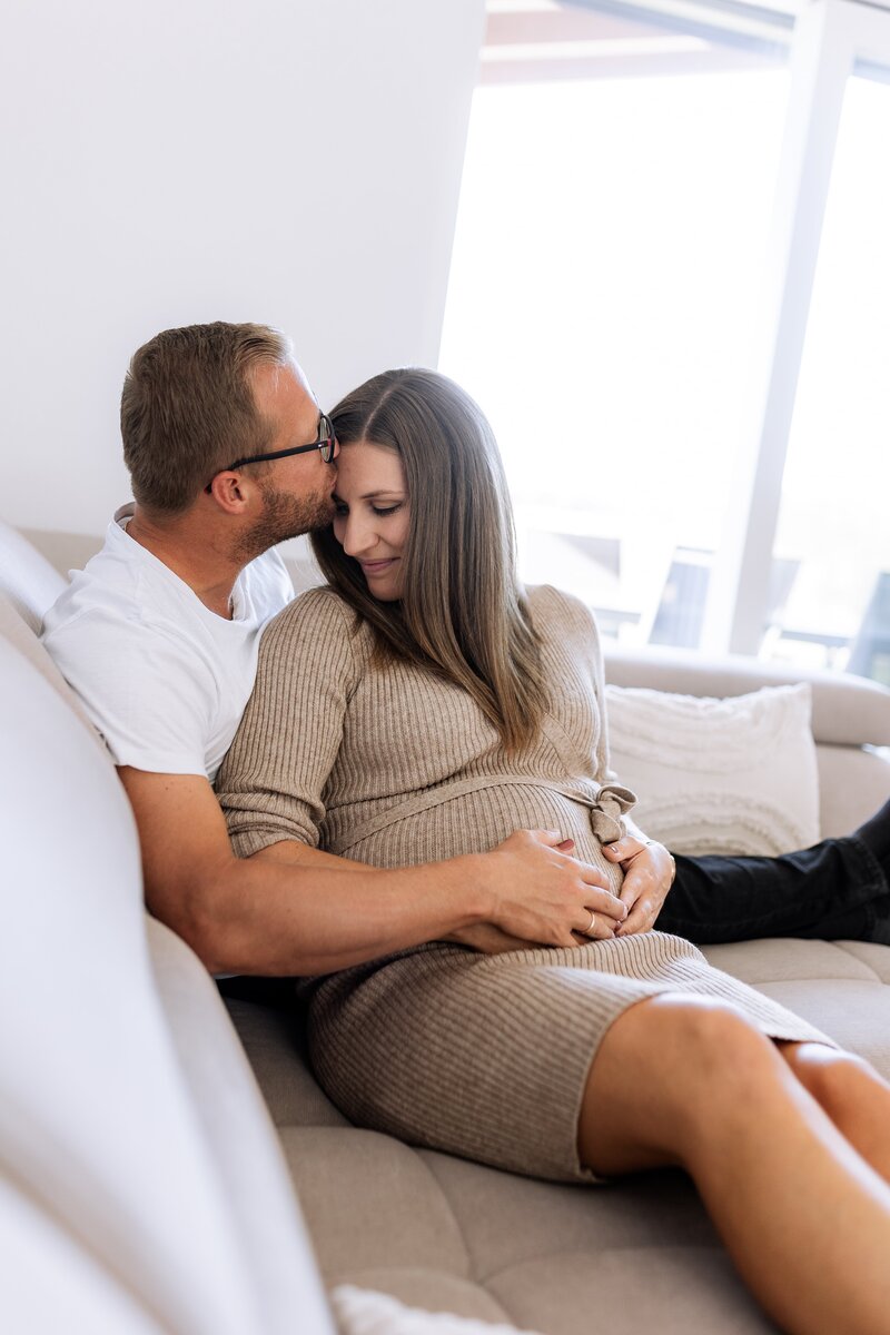 Paar sitzt auf Sofa und Mann kuesst den Kopf der Frau und haelt  ihren Babybauch