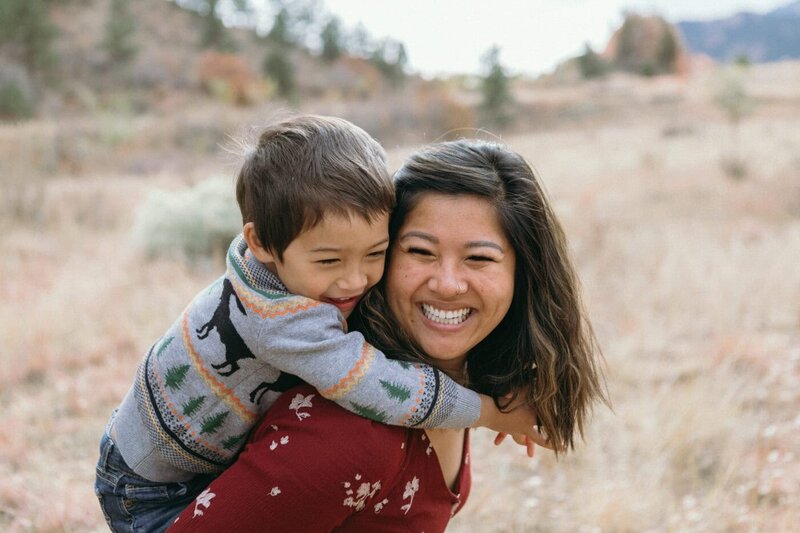 A toddler rides piggyback on his mom and makes her smile.