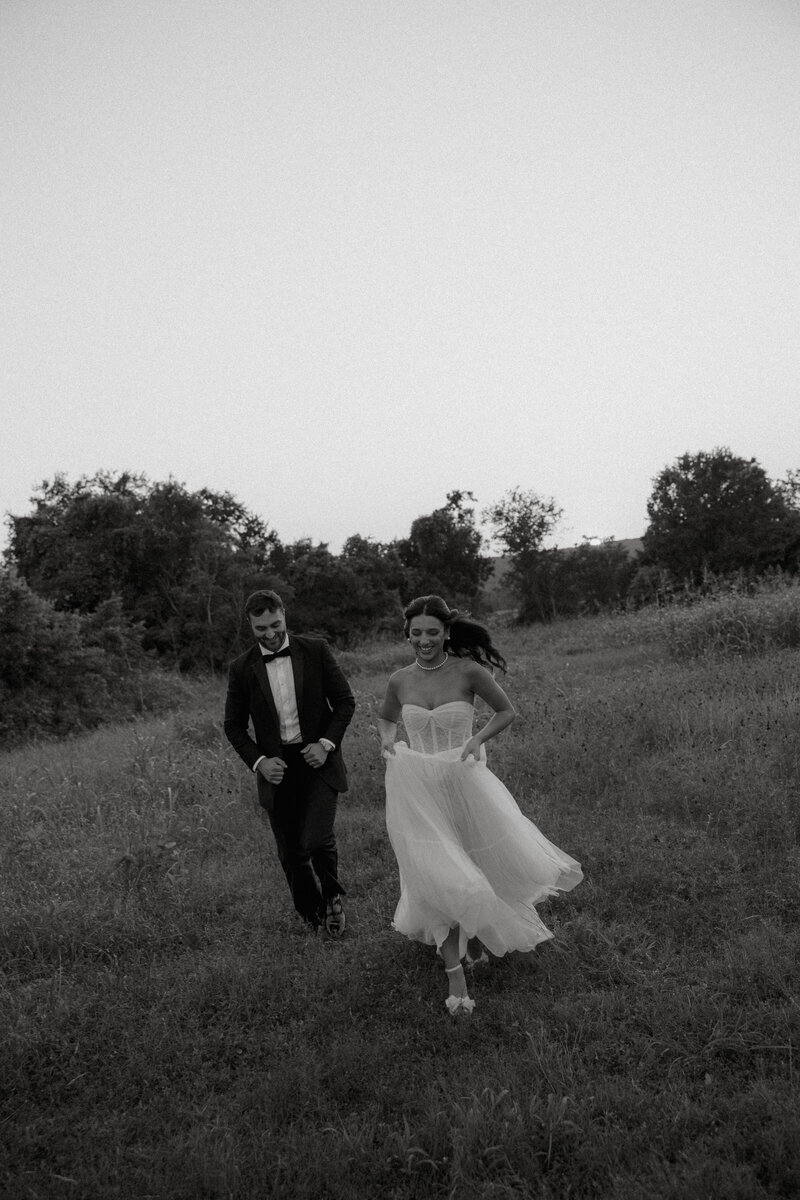 Black and white photo of bride and groom running through grass