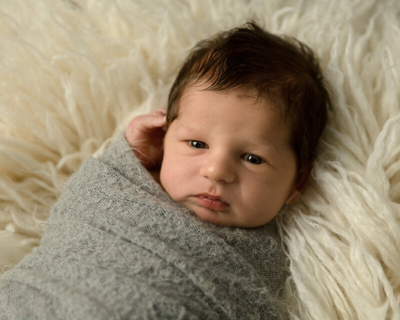 Newborn boy with a full head of dark hair laying awake on a white rug while wrapped in a gray wrap.