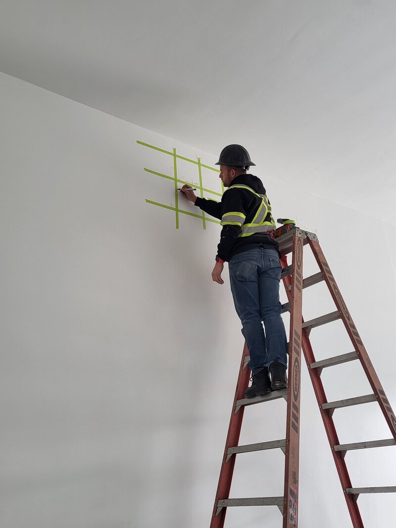 An operator on a ladder is marking object that the scanner found in the concrete by running lines of green tape in a grid along the wall