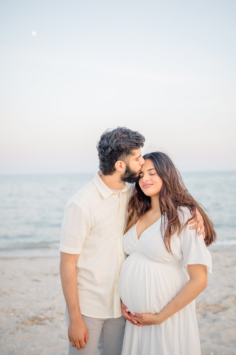A heartfelt maternity session by Oak & Lore Studios captures a husband gently kissing his pregnant wife's head as they stand together on a serene beach. The soft ocean breeze and golden light create a warm, intimate moment celebrating their growing family