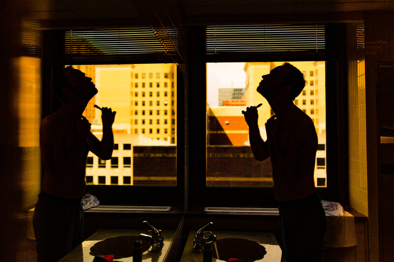 A groom shaving next to a window over looking  Downtown Toledo Ohio