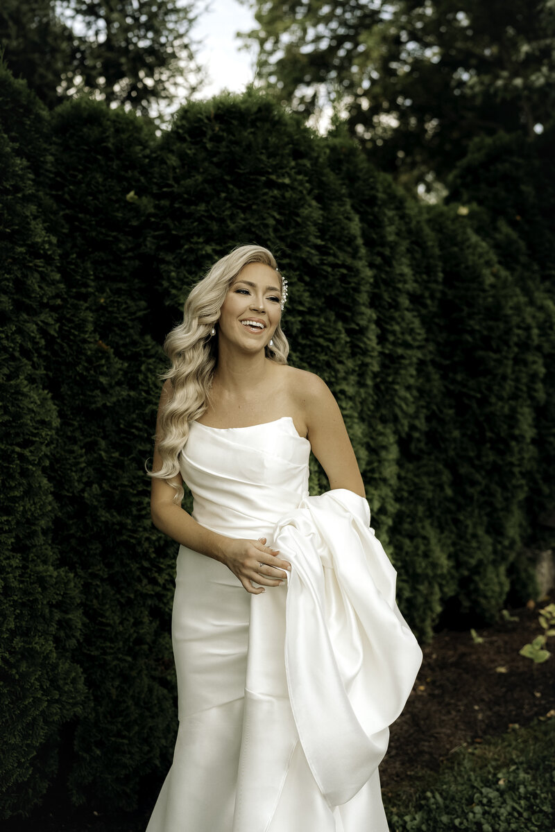 A bride standing in front of tall bushes smiling to the side while holding her dress with long wavy blonde curls.