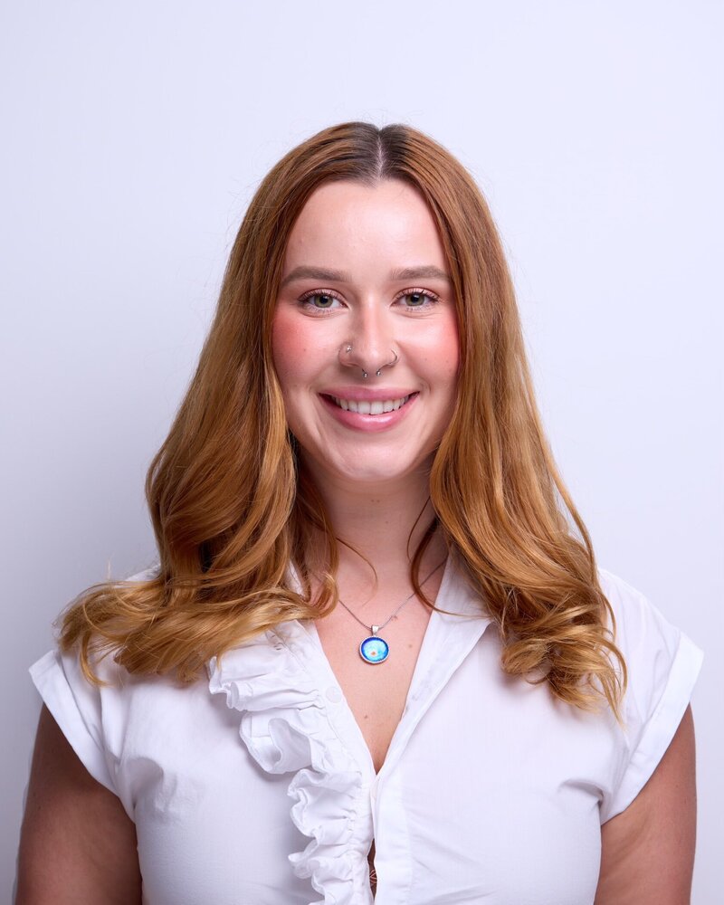Woman with long hair smiling. She has layered hair, blond or brown in color. The photo is a portrait shot indoors, showing her face, neck, and part of the wall behind her.