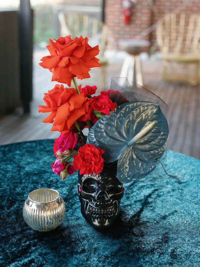 black skull vase with rave jeweled "makeup" and red roses, iridescent anthurium, and hot pink carnation arrangement sitting on a velvet teal linen hightop on the terrace at the Line Hotel DC