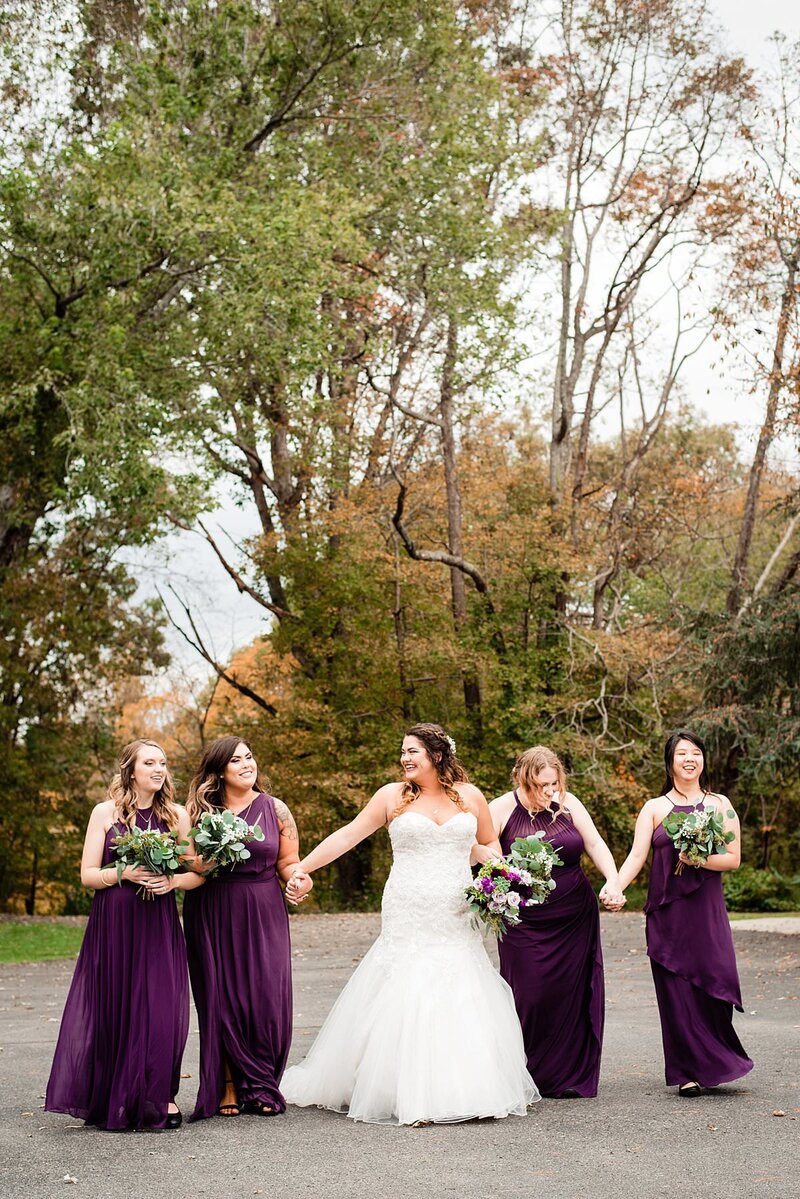 Bride with her 4 bridesmaids around her in an eggplant purple, they are holding hands and walking