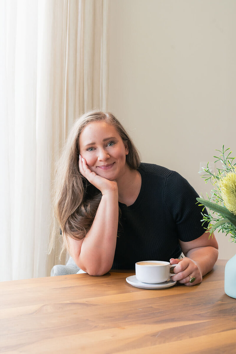 A woman sitting at a dining table, holding a coffee cup and smiling