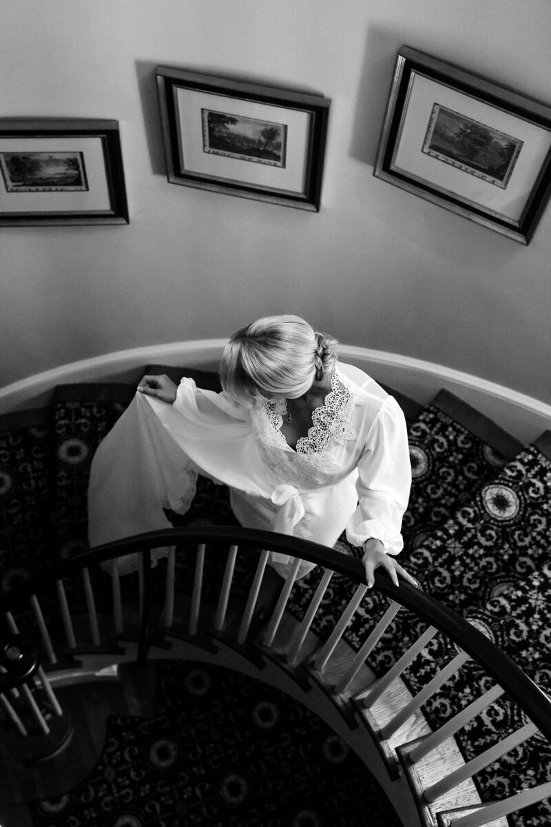 Brittany gracefully walks down the stairs at Boxwood Estate, preparing for her wedding day. Her hair is pulled back into an elegant bun, and she wears a delicate white lace robe. The soft, natural light illuminates the moment, highlighting her serene expression as she makes her way toward the ceremony. The elegant interior of the estate provides a beautiful backdrop, adding to the sense of anticipation and excitement.