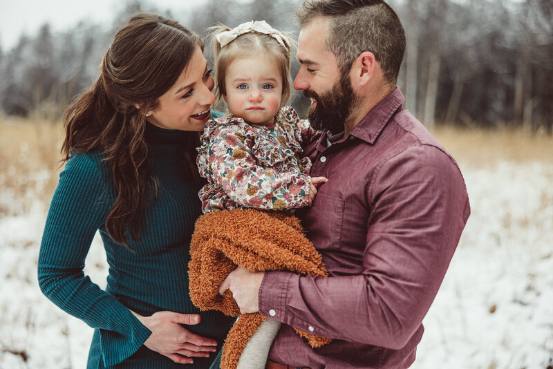Expecting family standing in the snow with parents looking at daughter who is in the middle being held my her father.
