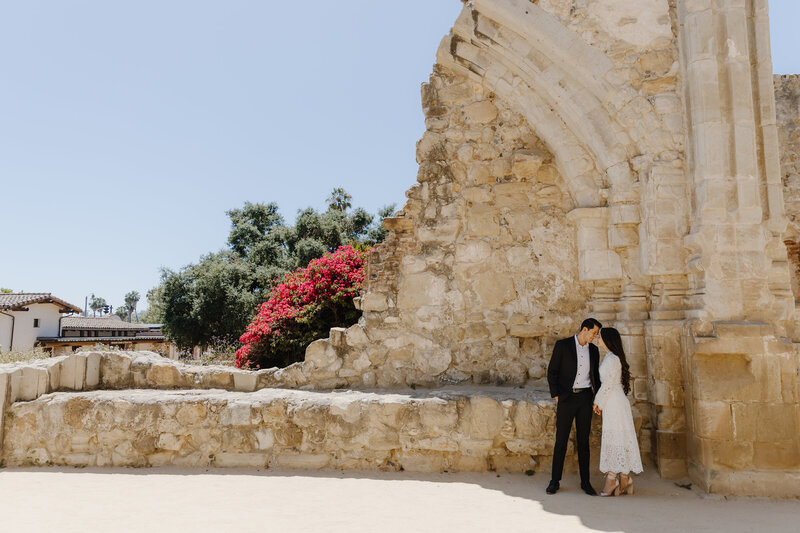 Couple kisses beside a sunlit stone wall and blooming bougainvillea at a Spanish-style mission.