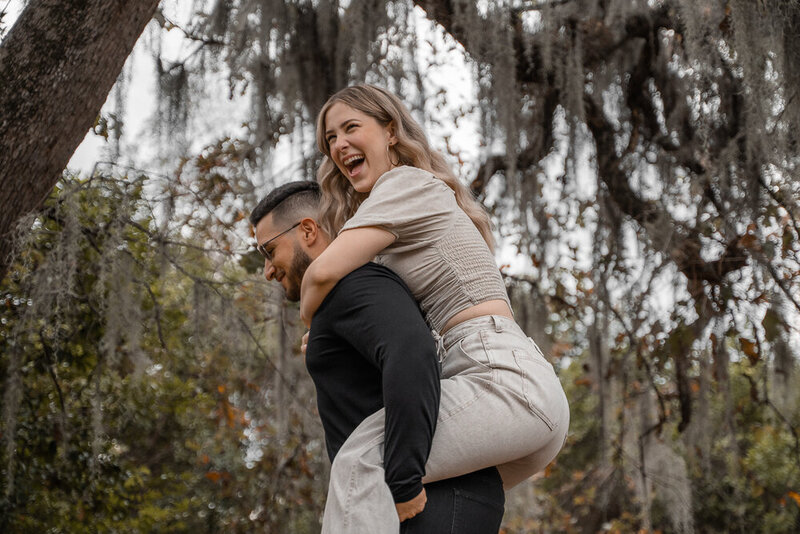 Engagement photoshoot of fiance on her fiances back and laughing with Spanish moss trees in the background.