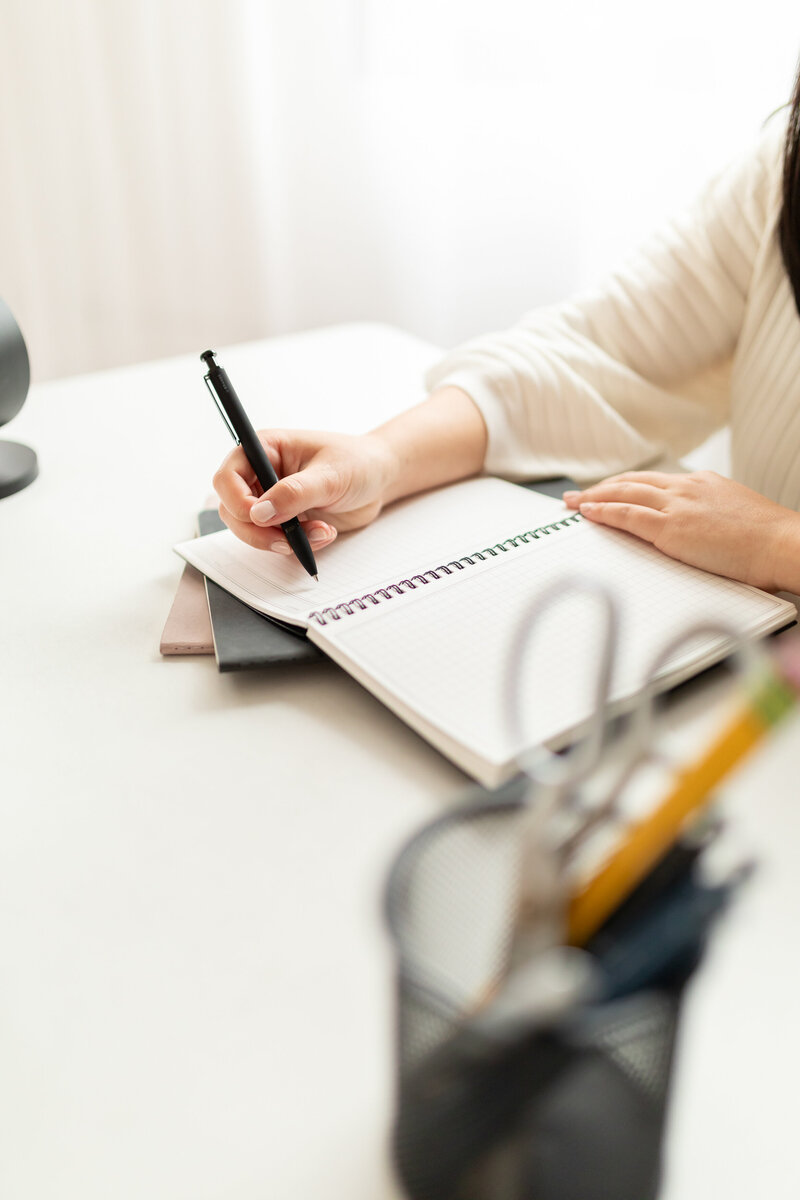 close up of a woman holding pen to paper planner with typical desk items around