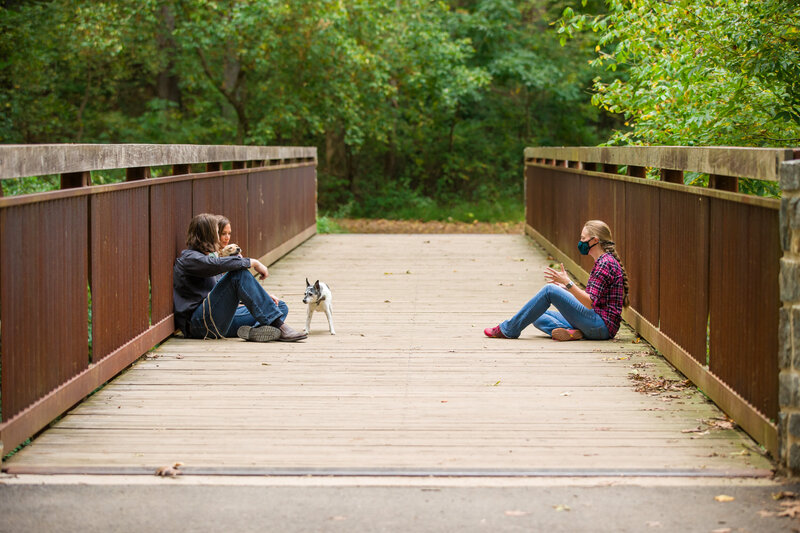 Jen Stitt sitting with an engaged couple on a bridge during an engagement session, sharing a relaxed and genuine moment while connecting with them to create a comfortable and natural photography experience.