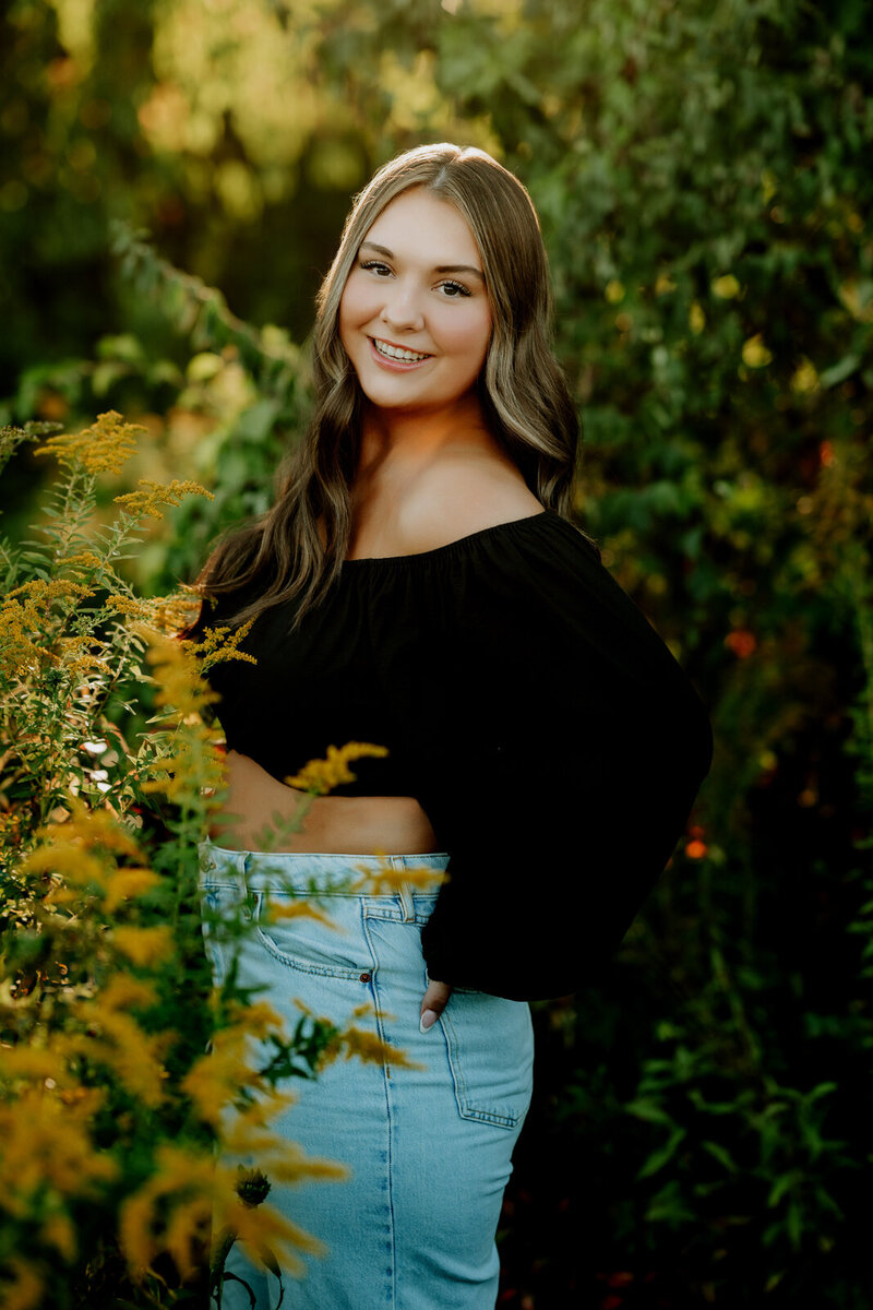 Girl wearing jeans and black top and yellow flowers posing for her high school senior photos. 