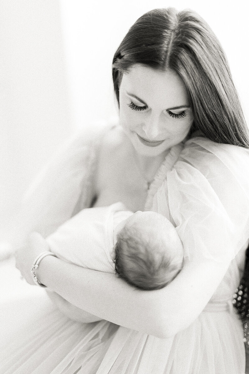 Close up light filled black and white photo of a mother dressed in a tulle gown while she holds her newborn baby boy and looks down at him.