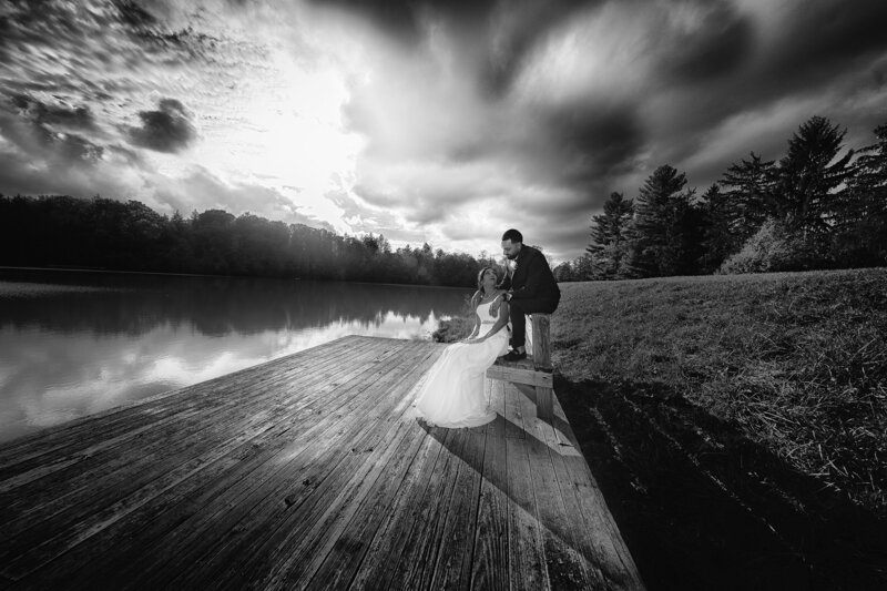 A black-and-white wedding portrait of a couple on a wooden dock beside a still lake