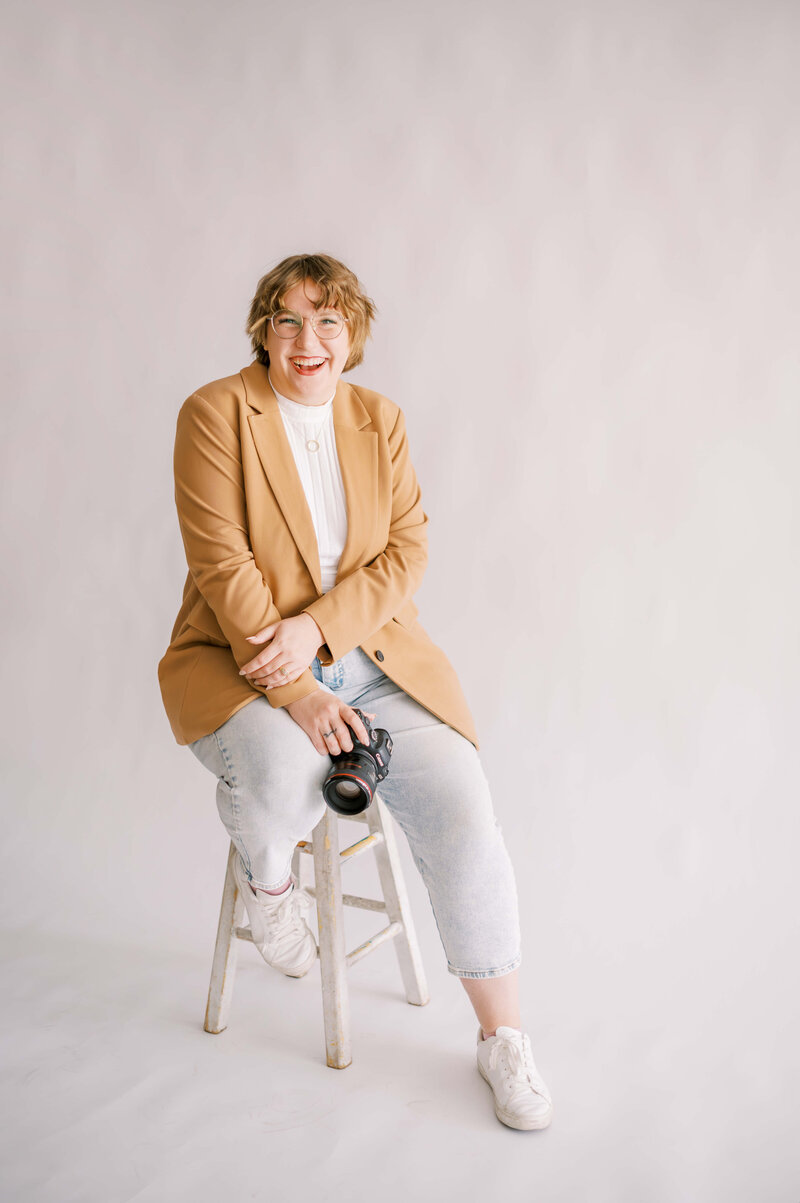 full body portrait of orange county wedding photographer Sam sitting on a stool in front of a white backdrop smiling at the camera