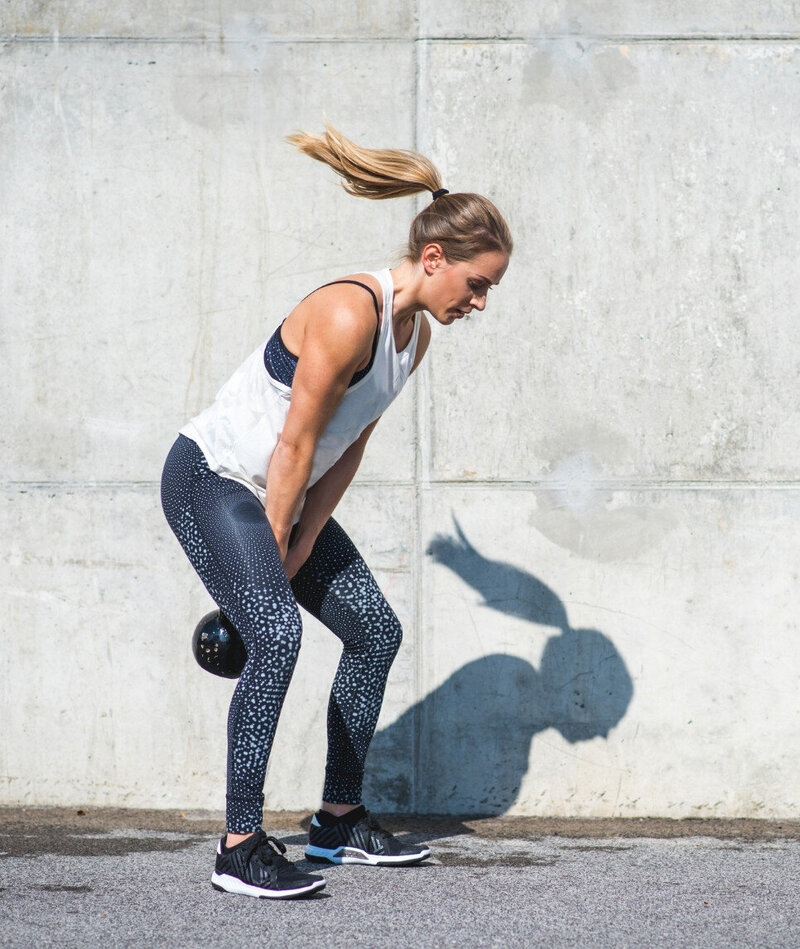 Woman performing a kettlebell swing outdoors against a concrete wall, representing strength and movement in midlife with Rebells.