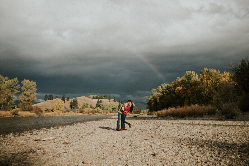 A man and woman kiss as she pops her leg in the air in front of a lake and a rainbow in the sky