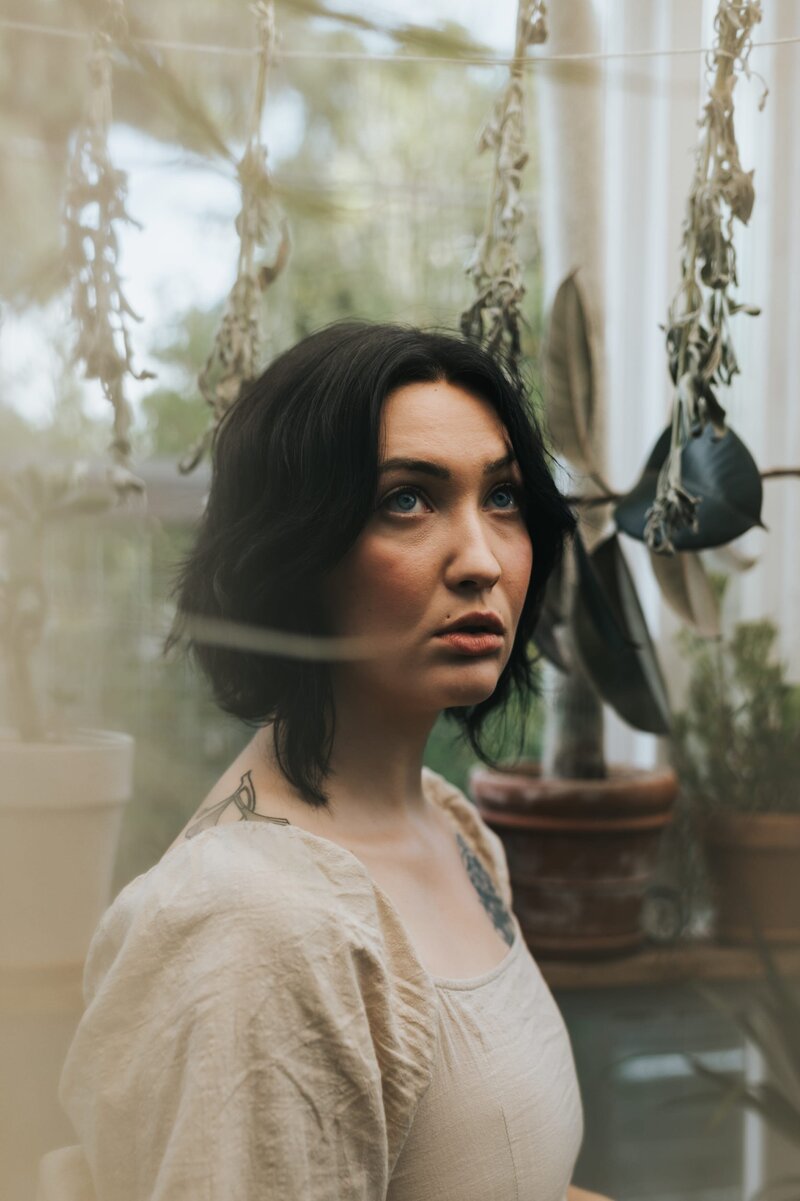 Woman with short dark hair wears a tan dress and looks up as she stands in a greenhouse surrounded by herbs and plants.