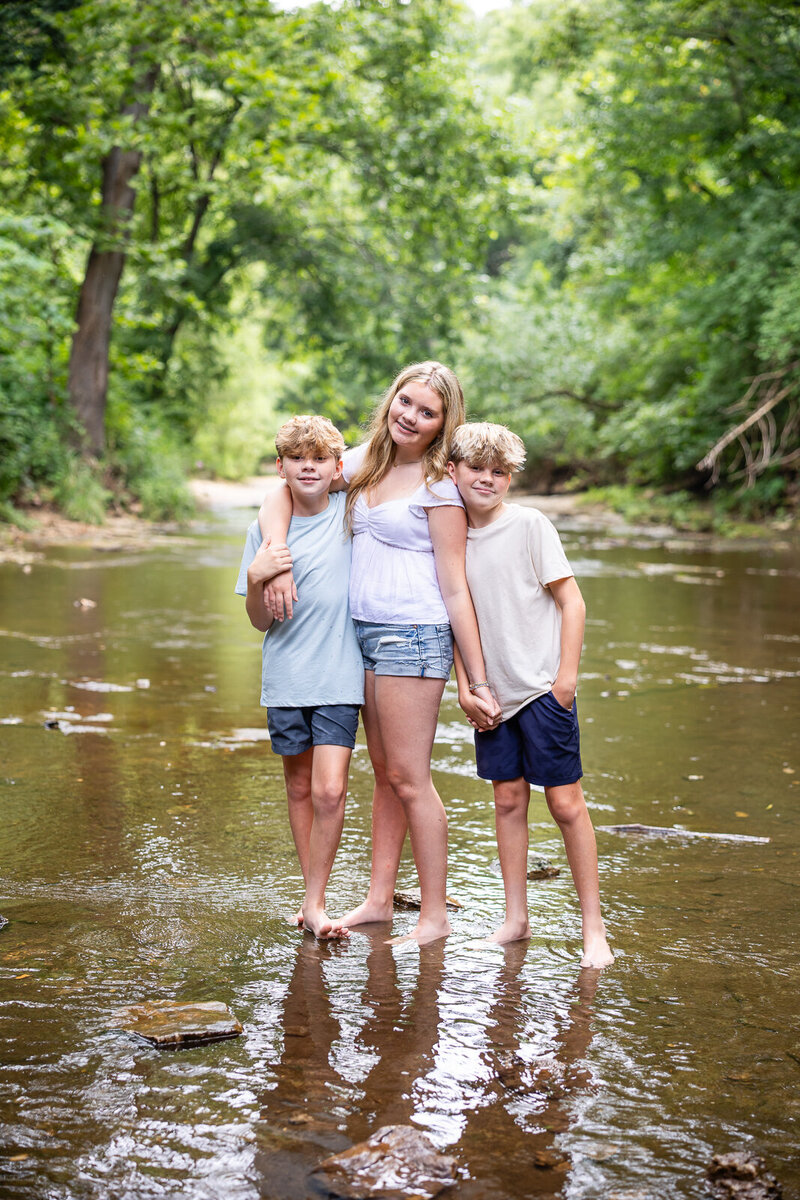 Big sister with her twin brothers standing in a creek smiling at the camera in Hillsboro, Missouri. 