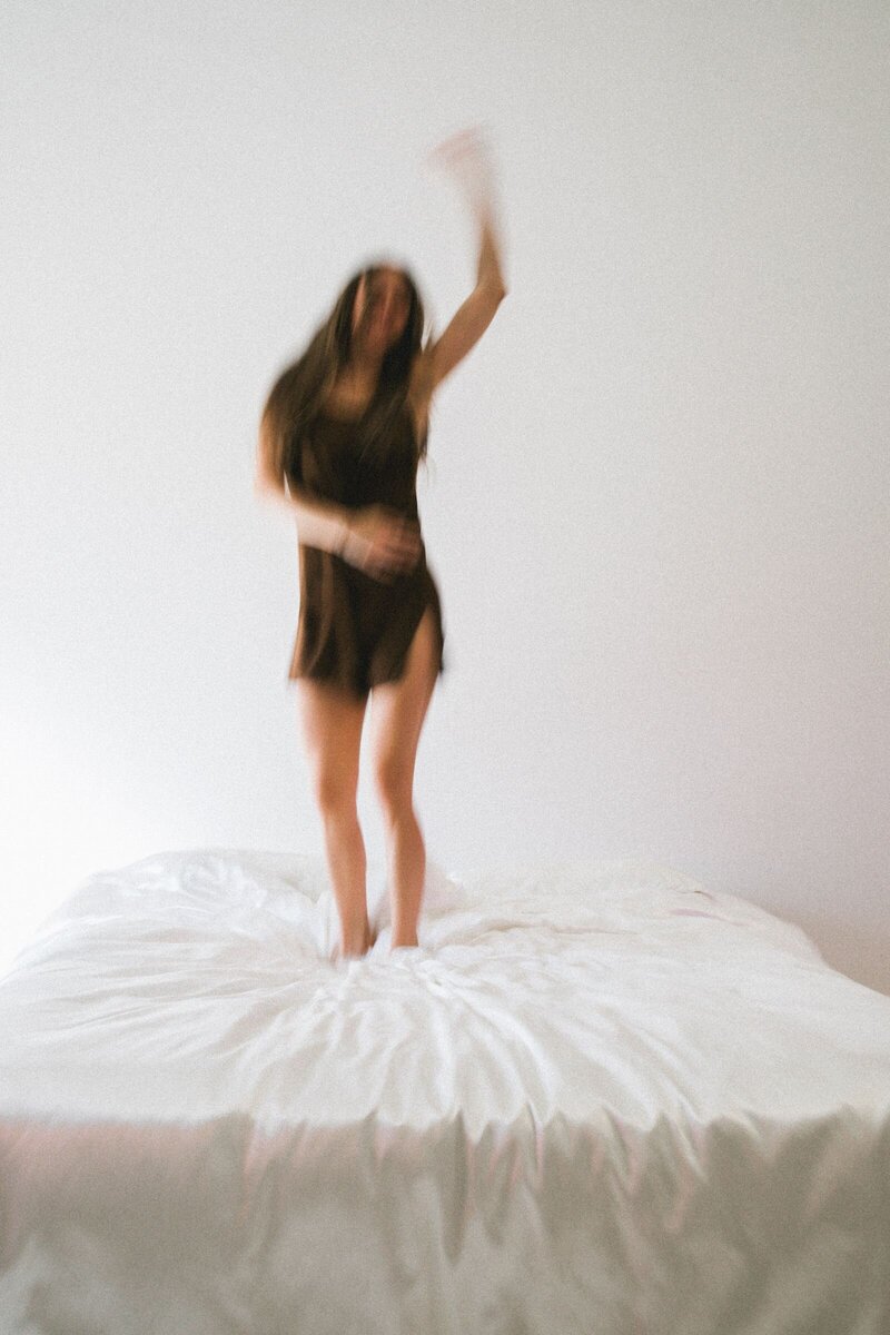 A woman with long hair, wearing a brown silk slip, joyfully jumps on a bed covered in white silk sheets. Her arm is raised in the air, capturing a moment of carefree elegance and energy.