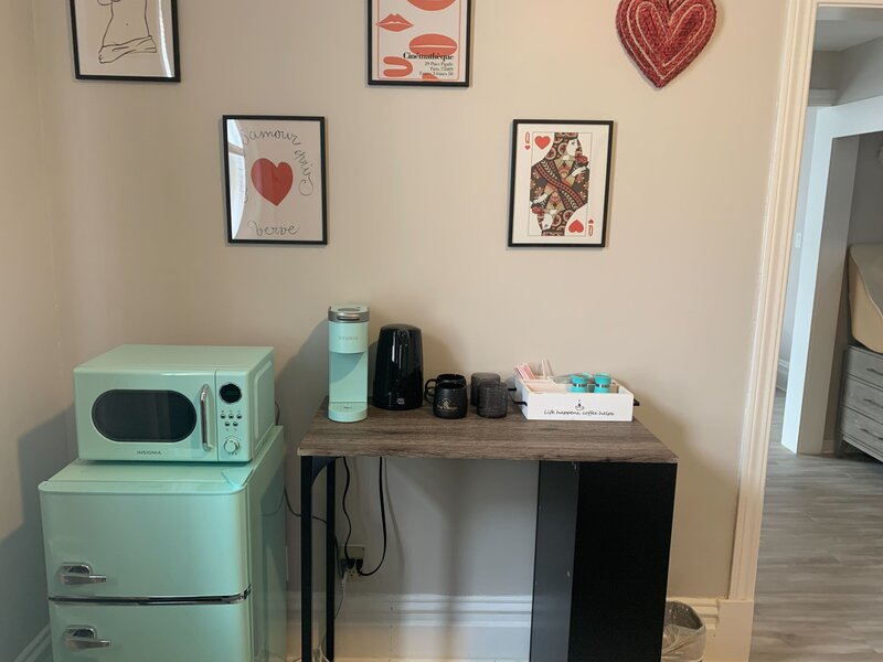 A close-up of one of the room's kitchenette. The microwave sits on the minifridge, both are mint green. A mint green coffee maker is on the table next to them along with an electric kettle, two mugs, and a box with assorted tea and coffee. Heart-themed art is on the wall.