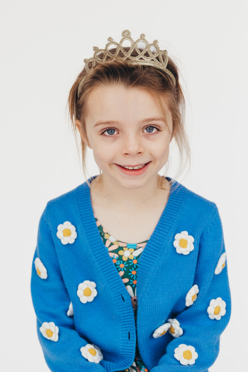 A young girl wears a colorful sweater and a cute gold crown during her portrait in front of a clean white background