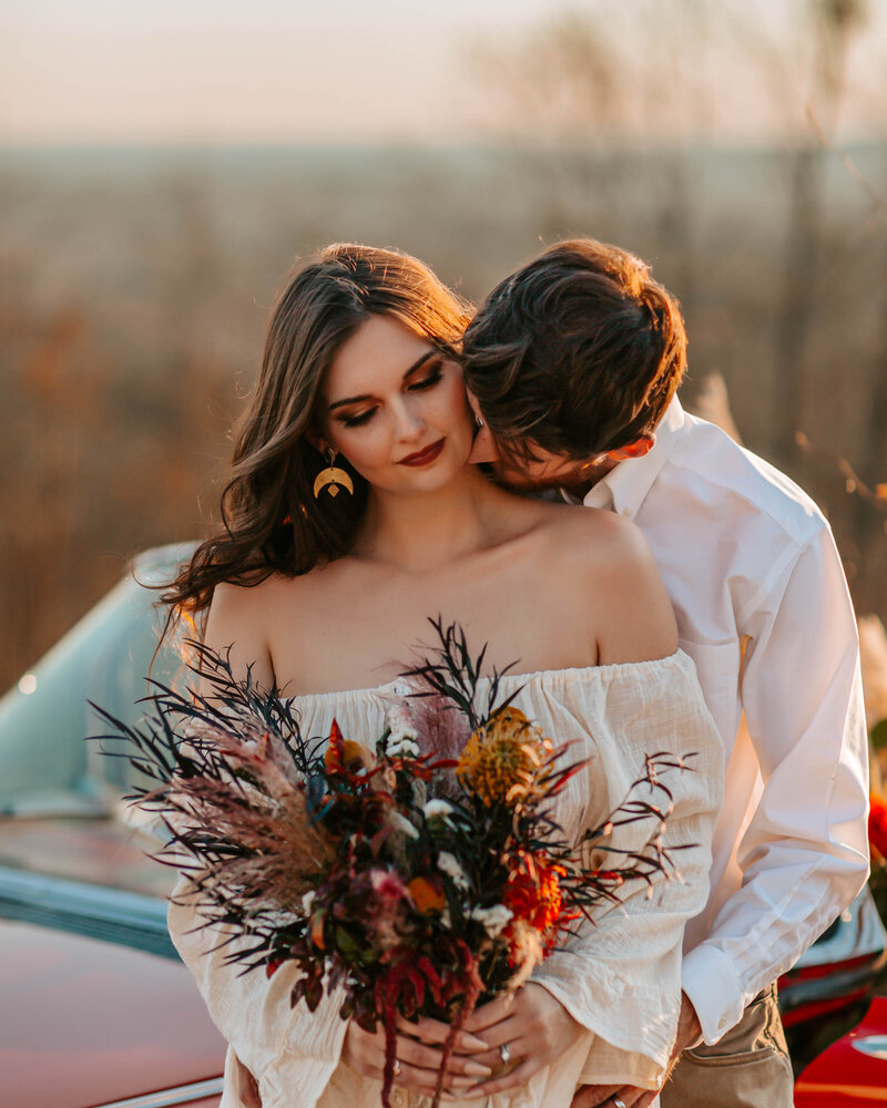 Wife standing with a bouquet of flowers while husband kisses her neck.