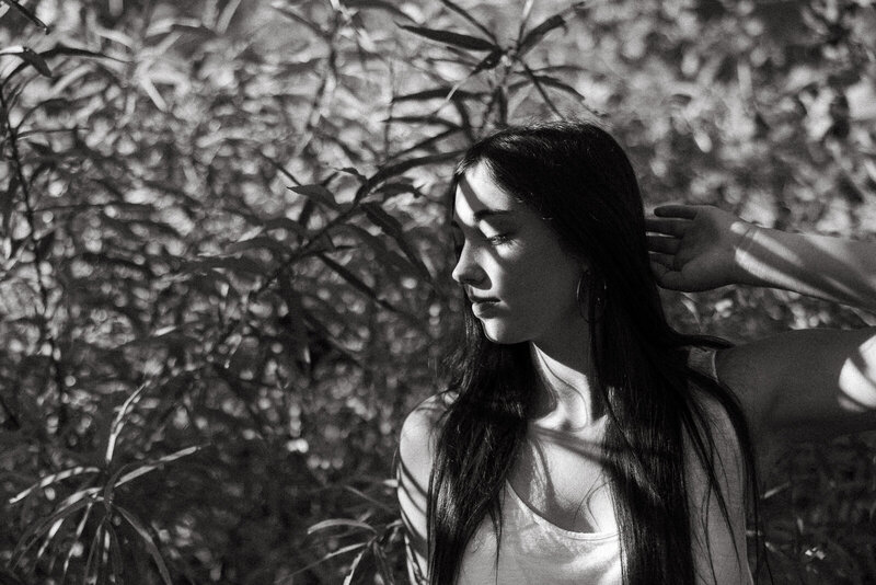 Black and white senior portrait of a girl standing near tall foliage with the shadow of the leaves laying across her face. 