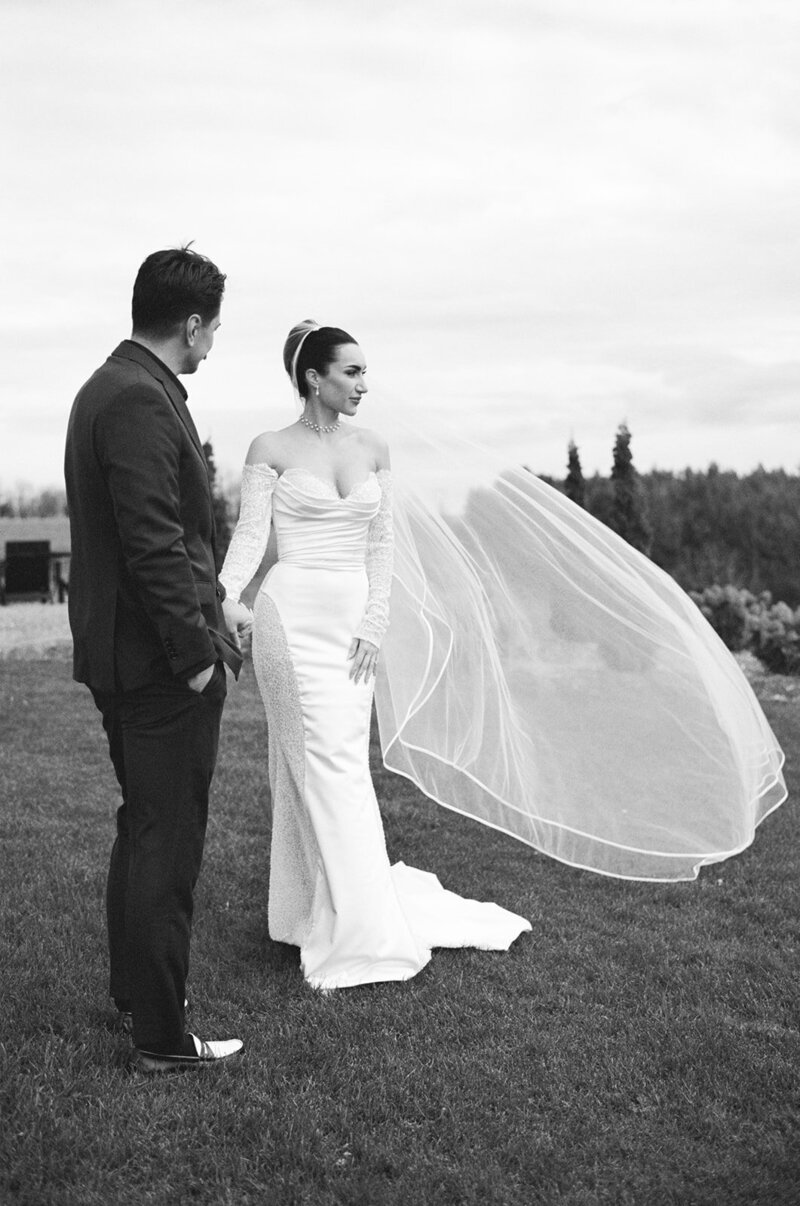 Bride in a long, white gown with a flowing veil stands with groom in a suit, holding hands outdoors. The enchanting scene captures the magic of wedding film photography, perfect for immortalizing love in Toronto's picturesque landscapes.