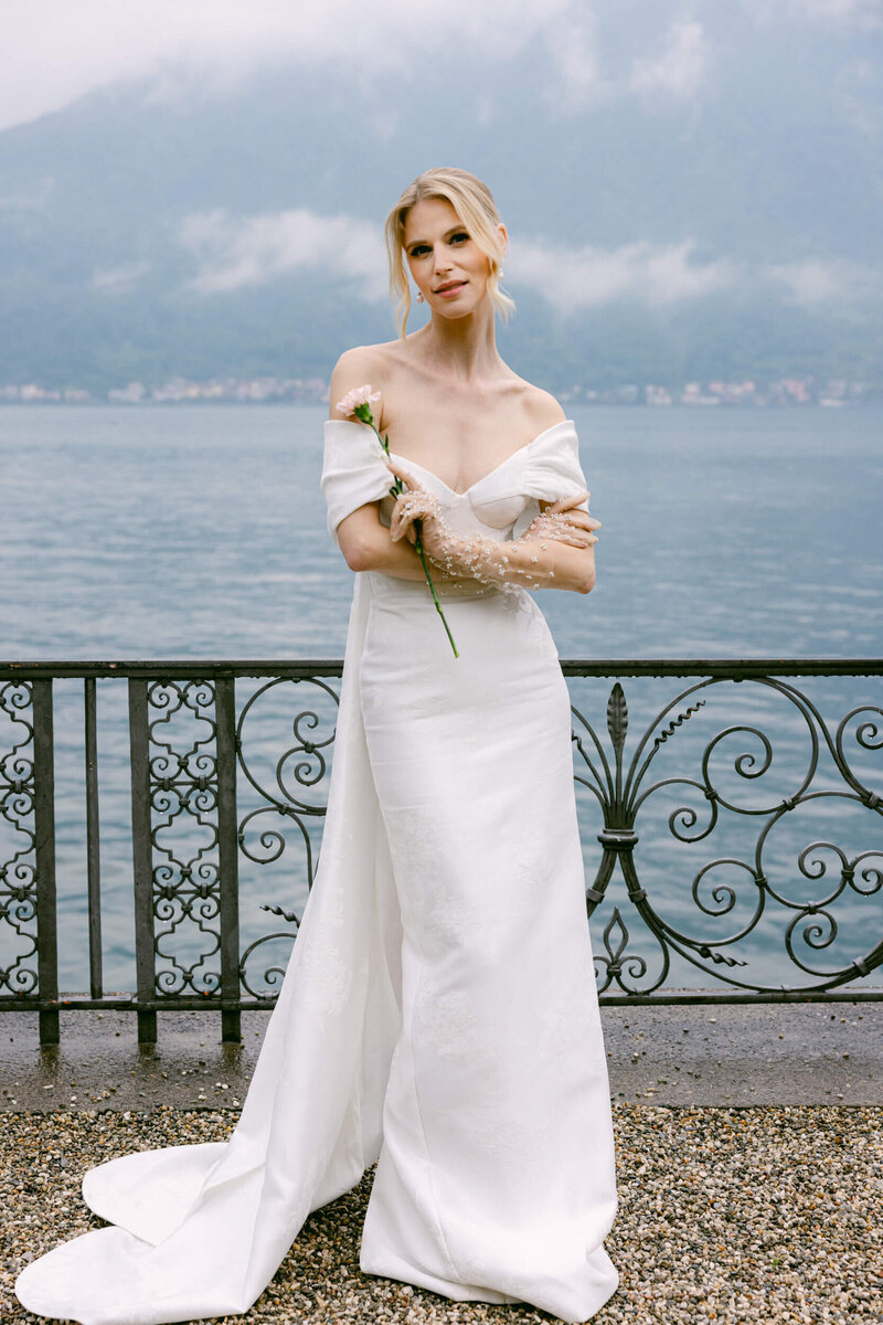 A wedding photo of a bride in a  white dress holding a rose on the shores of lake como in italy at villa balbiano