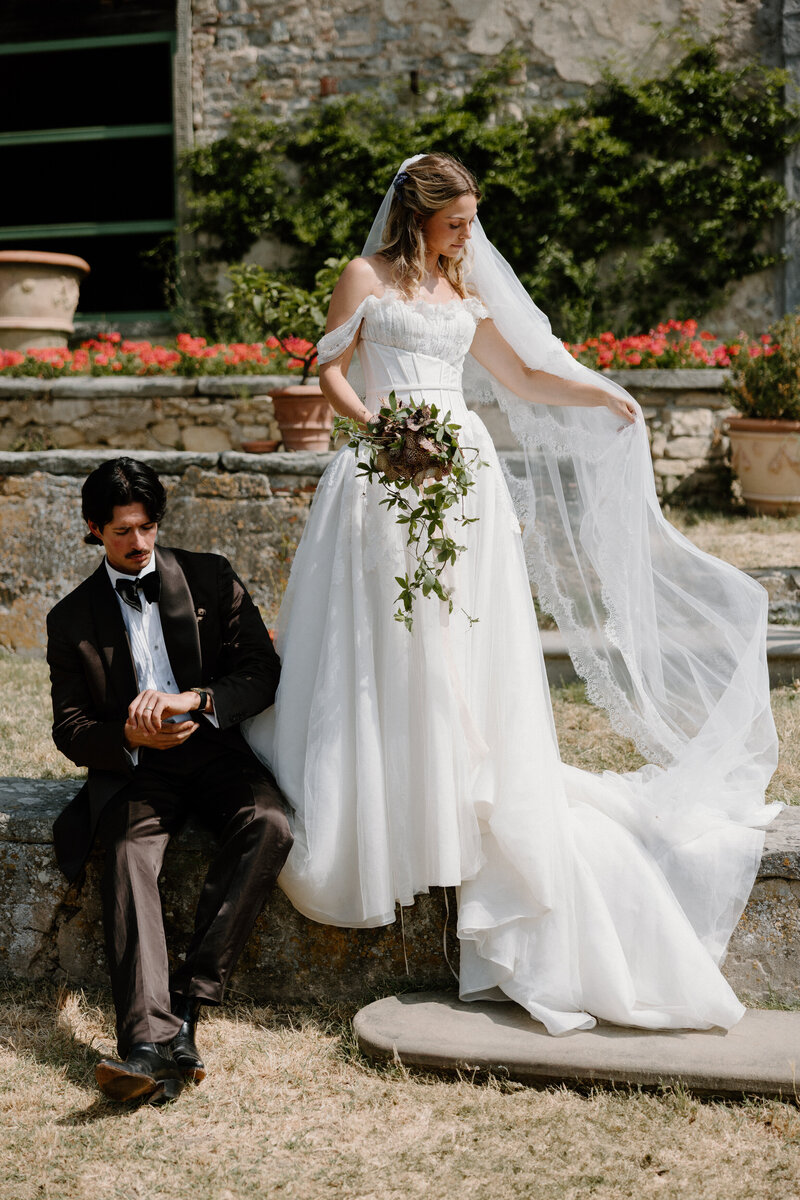 A person in a wedding dress adjusting their veil with their partner sitting next to them 