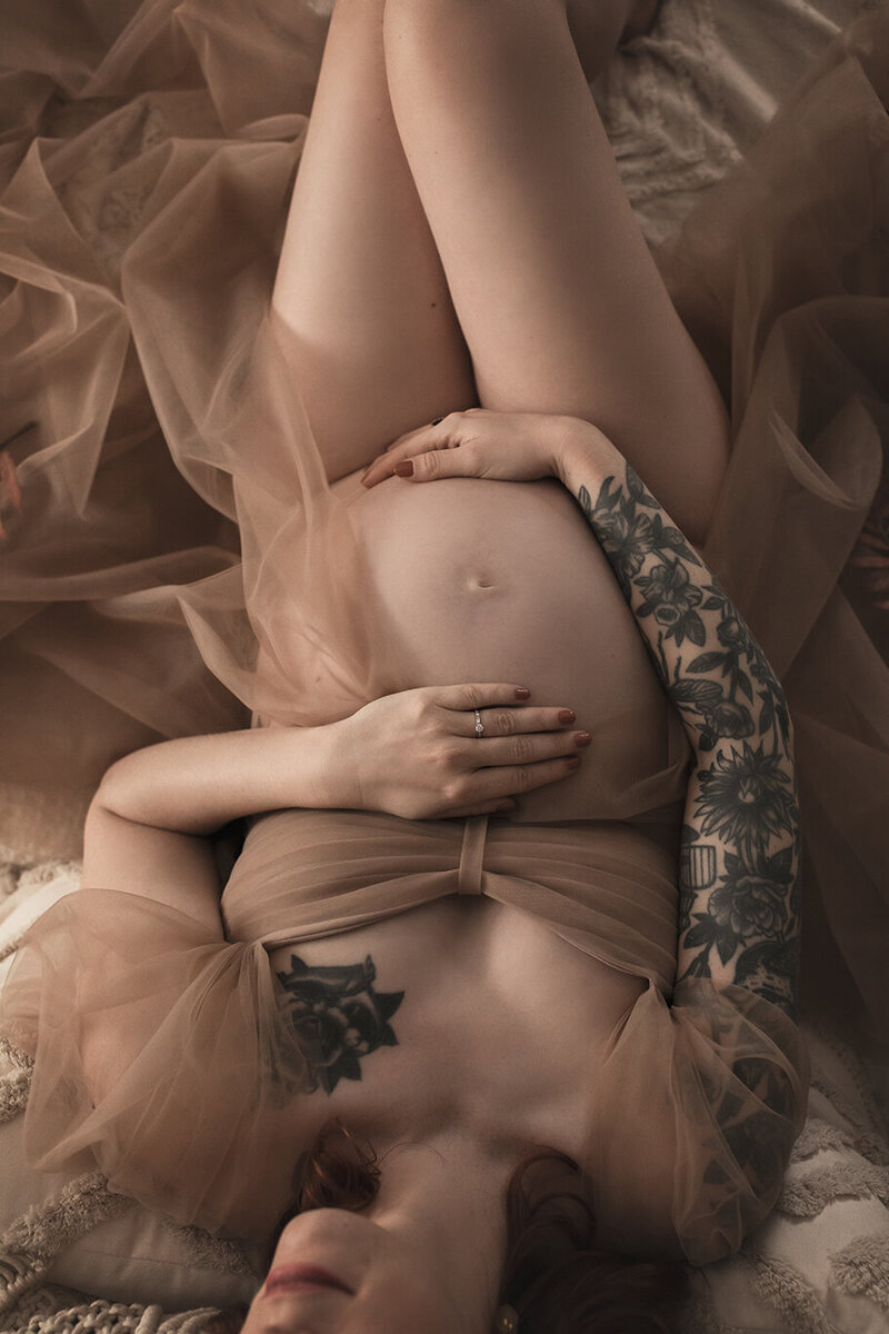 Femme enceinte allongée en robe nude transparente, mains posées sur son ventre, lumière douce, tatouages visibles sur le bras.