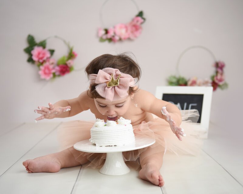 A baby girl in a pink ruffled dress, holding golden "ONE" letters with a flower headband, sitting on a white wooden floor during her cake smash photoshoot.
