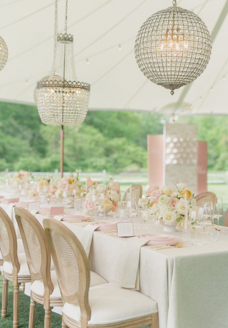 stunning tablescape at tented spring wedding. Multiple chandeliers and light wood french chairs with cream linens and pink plates. Floral centerpieces featuring pink, cream and yellow florals. a luxurious wedding.