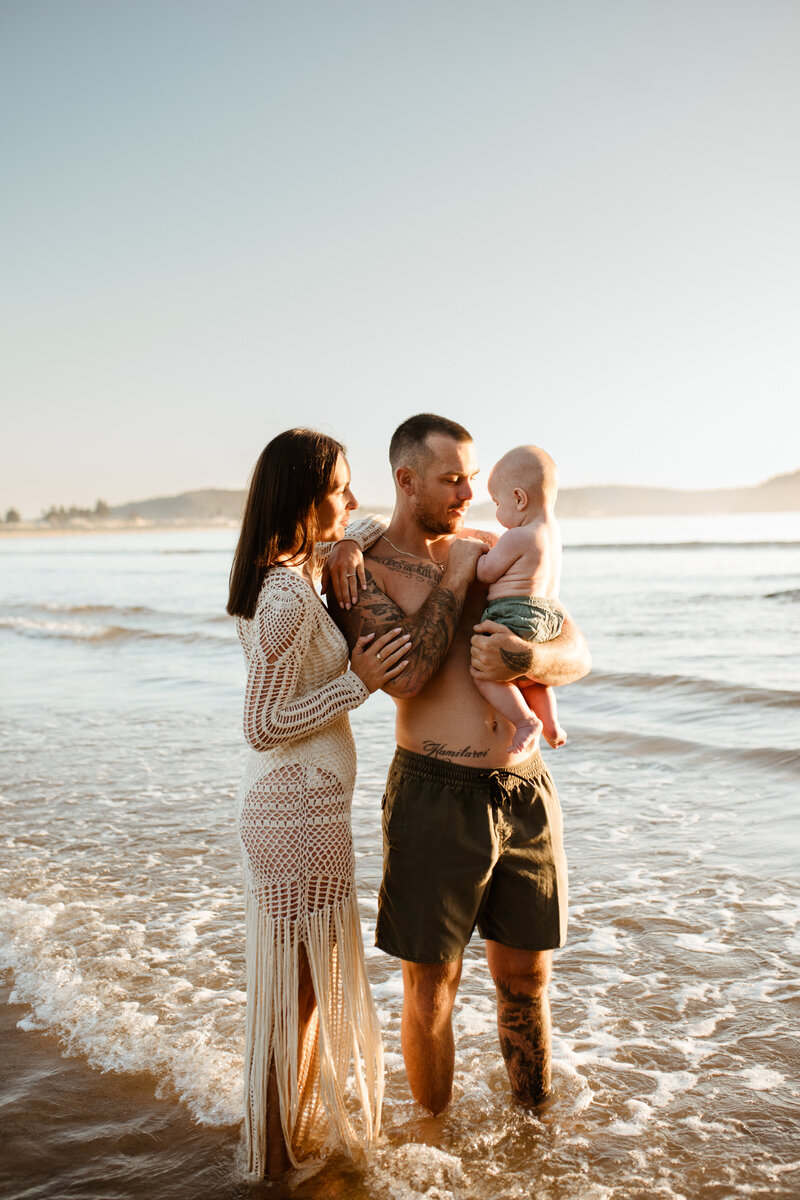 A family of three stands in shallow ocean water at the beach. The man is holding a baby, and the woman is standing next to them, both adults smiling at the child.